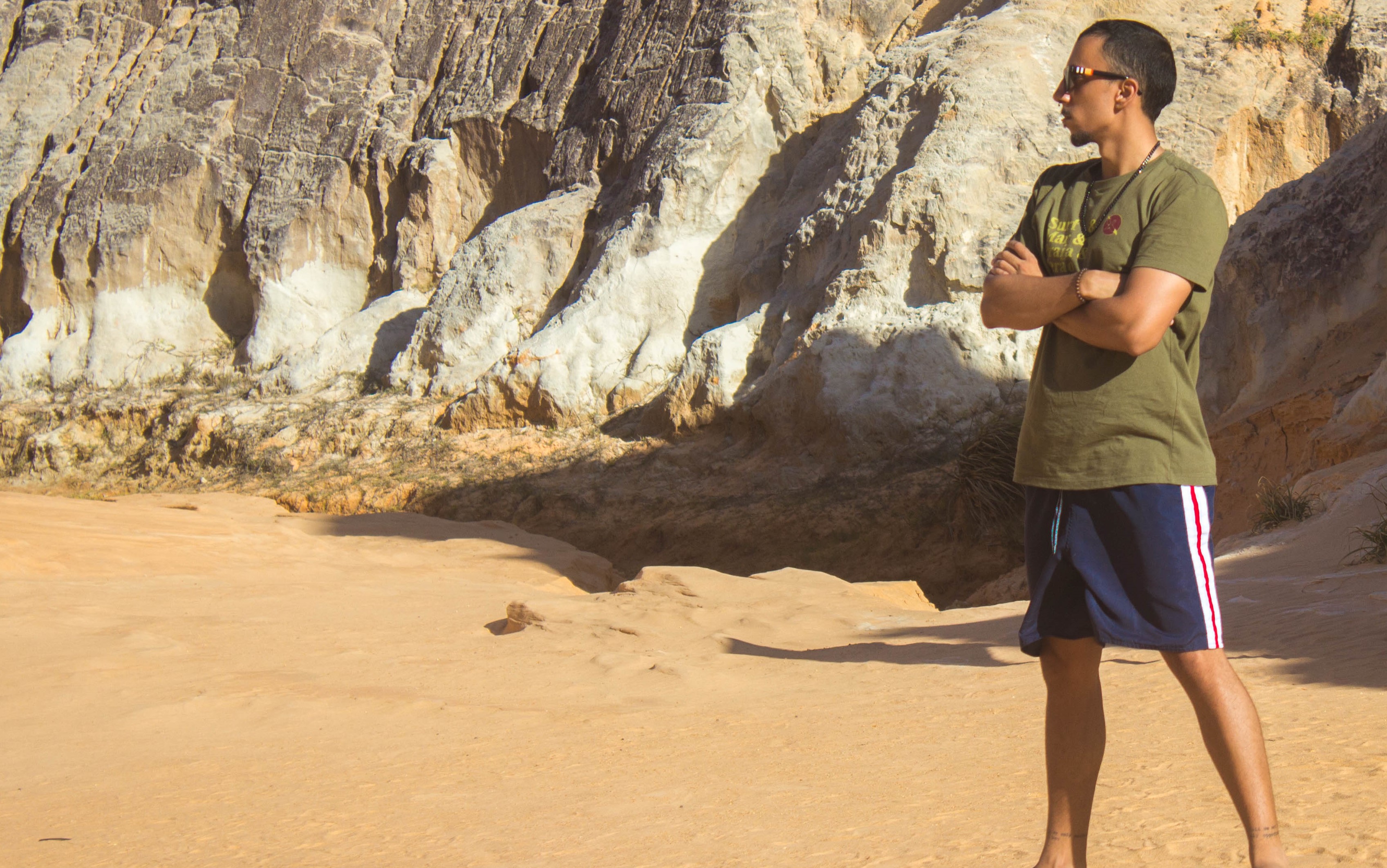 Jericoacoara, Ceará, Brazil Yuri on the right, standing sideways, looking left with his arms crossed. The entire ground is sand, the background is a rock all and the sun is at 45 degrees creating large shadows.