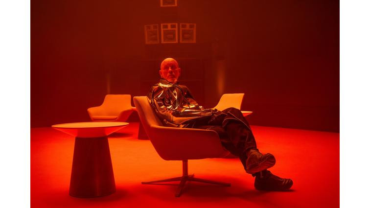 A man in a metallic coat and black trousers sitting in a chair next to a table in a room bathed in red light.