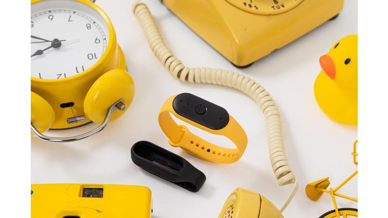 None Side angle of Bee wearable surrounded by yellow objects, including a yellow alarm clock, yellow rotary phone, rubber duck, and bicycle. To its lower left, you can see the black pin attachment.