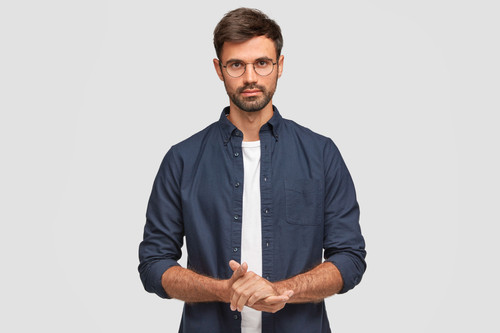 Waist up portrait of handsome serious unshaven male keeps hands together, dressed in dark blue shirt.jpg