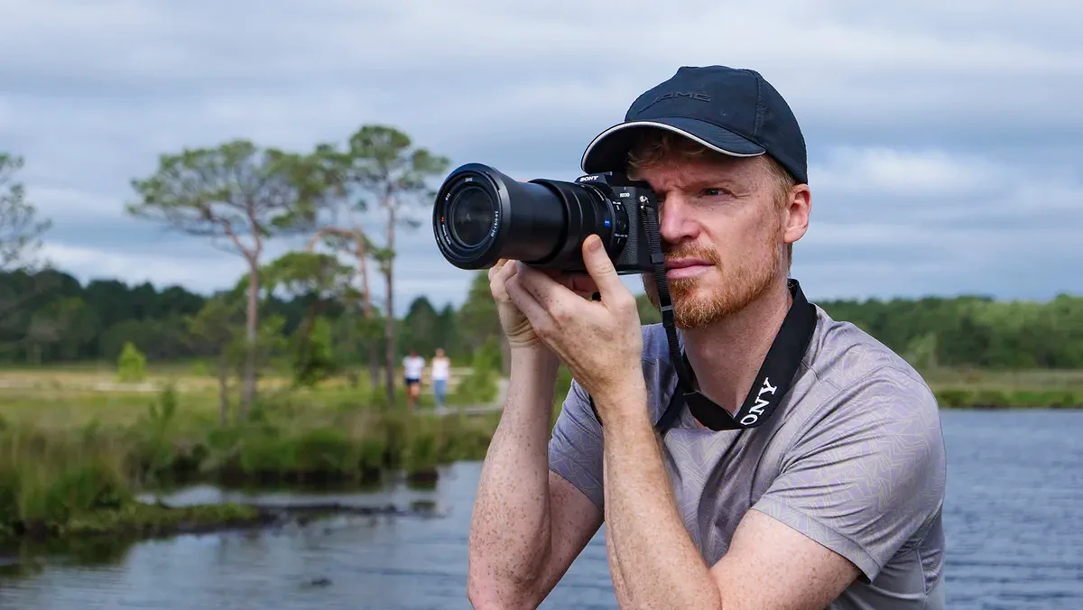Photographer and tech journalist Tim Coleman holding the Sony RX10 V bridge camera up to his eye in a nature reserve, with dramatic clouds behind him