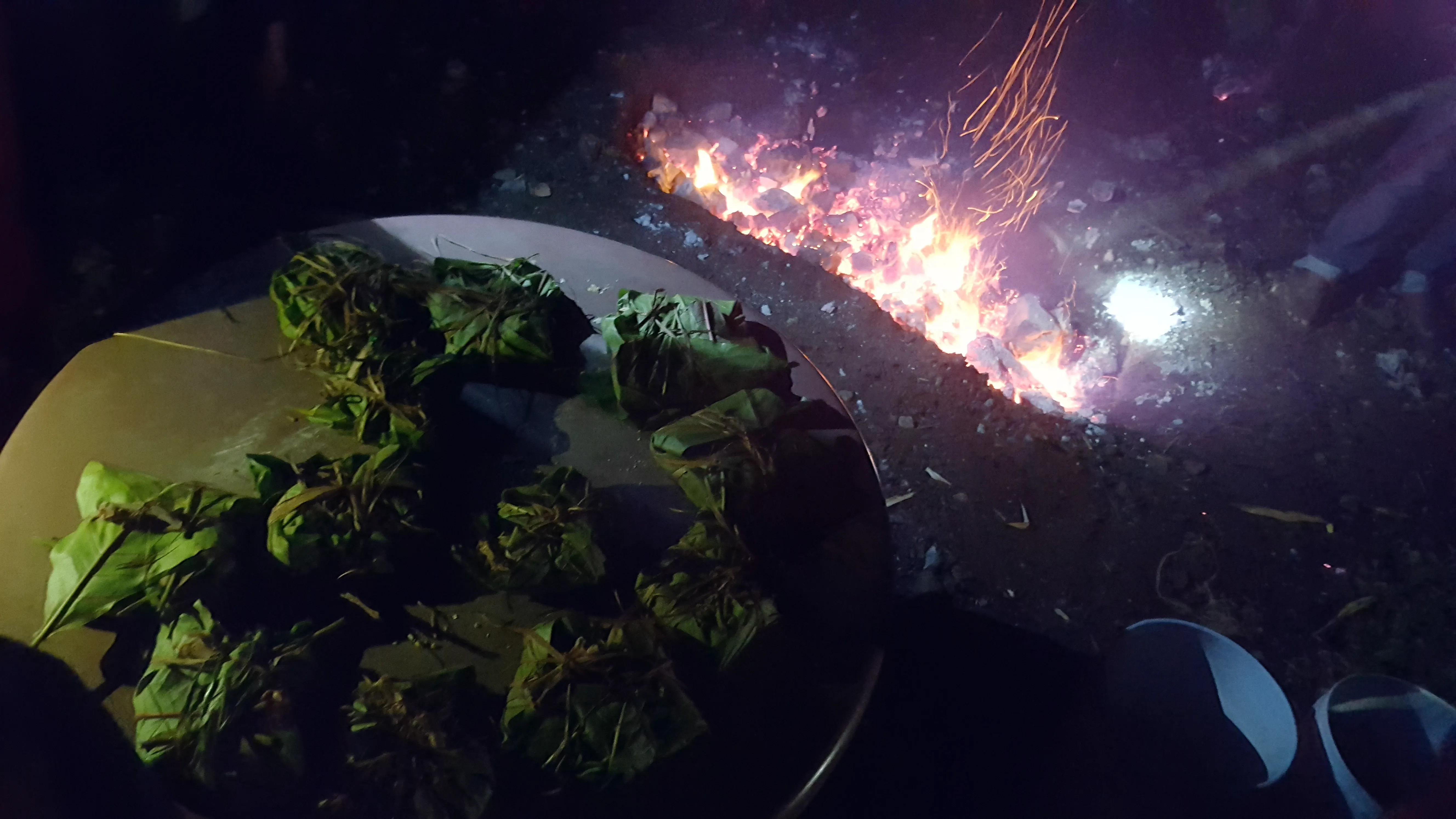 Mesa llena de pibes ya envueltos en hojas listos para meterse al horno bajo tierra con brasas que se ve al fondo