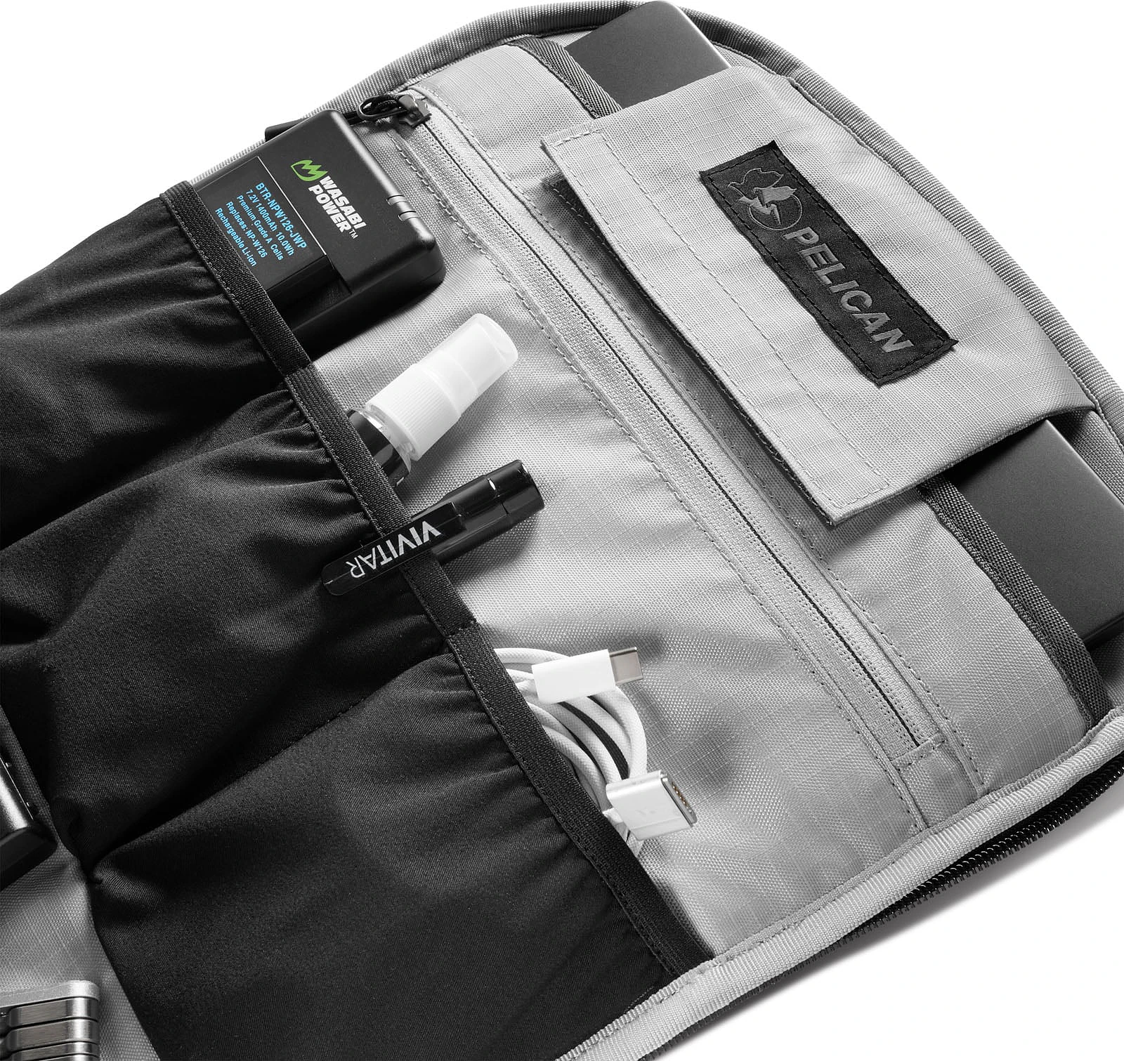An open Pelican bag interior featuring a black battery, a small spray bottle, a Vivitar pen, and a white charging cable stored in black fabric pockets.