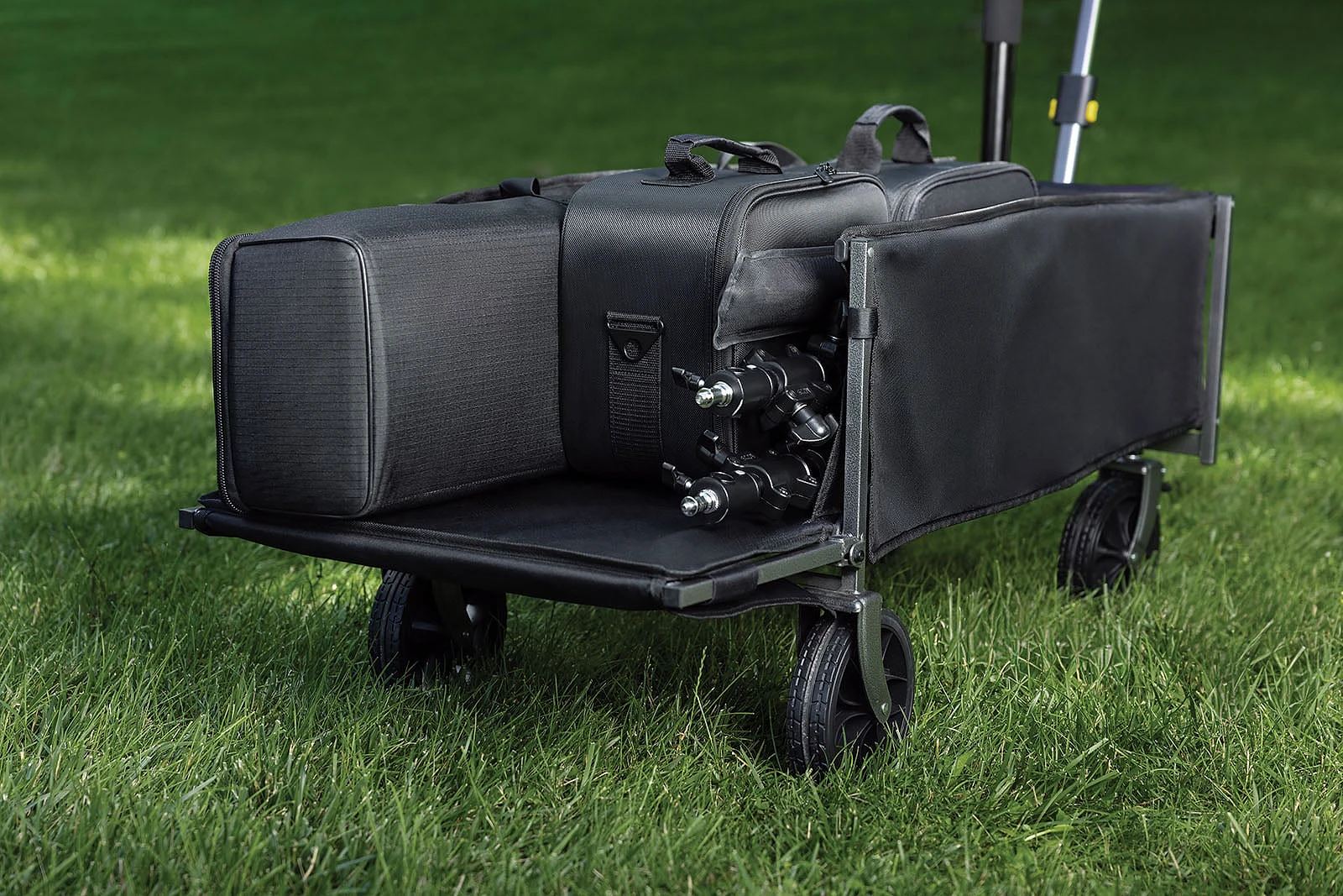 A black collapsible utility wagon sits on green grass, carrying several black equipment bags and a tripod head.