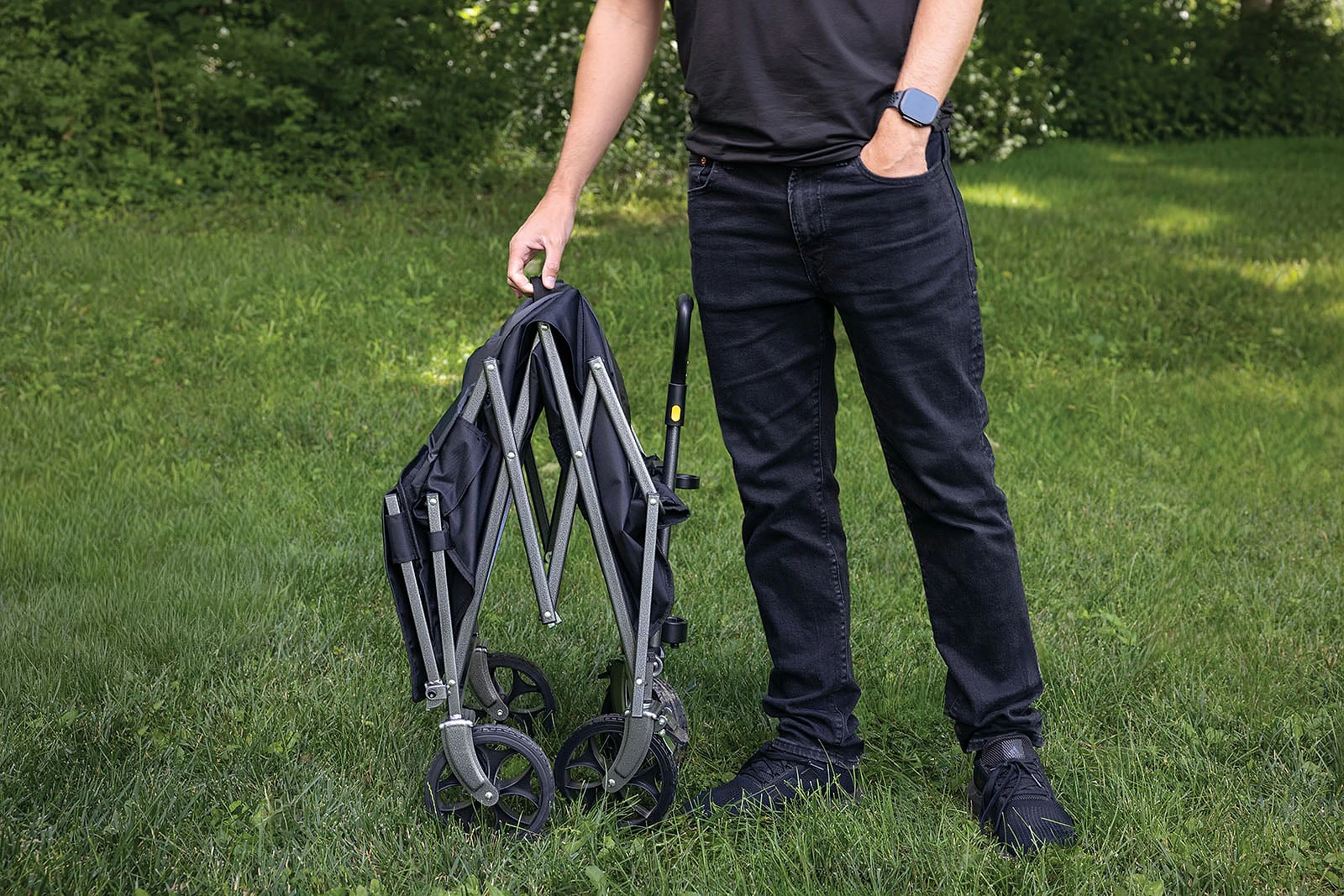 A person in a black t-shirt and jeans stands in a grassy field holding a folded black collapsible utility wagon.