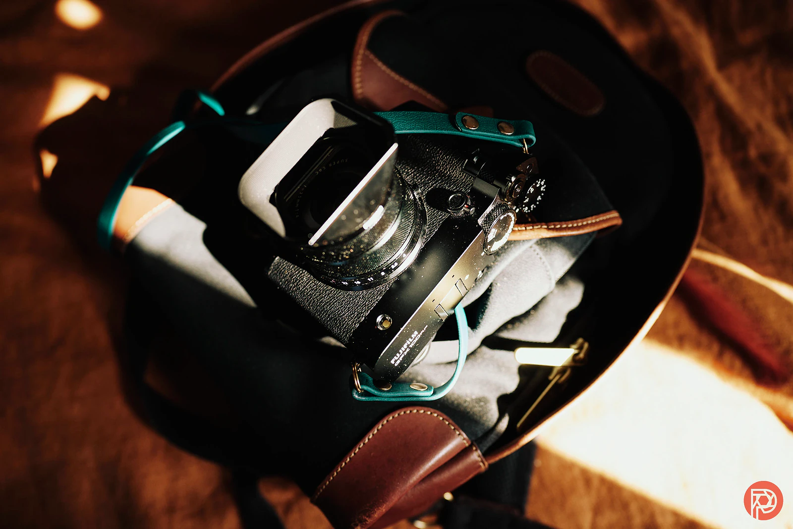 A black camera with a teal strap sits on top of a brown and black bag in warm sunlight.