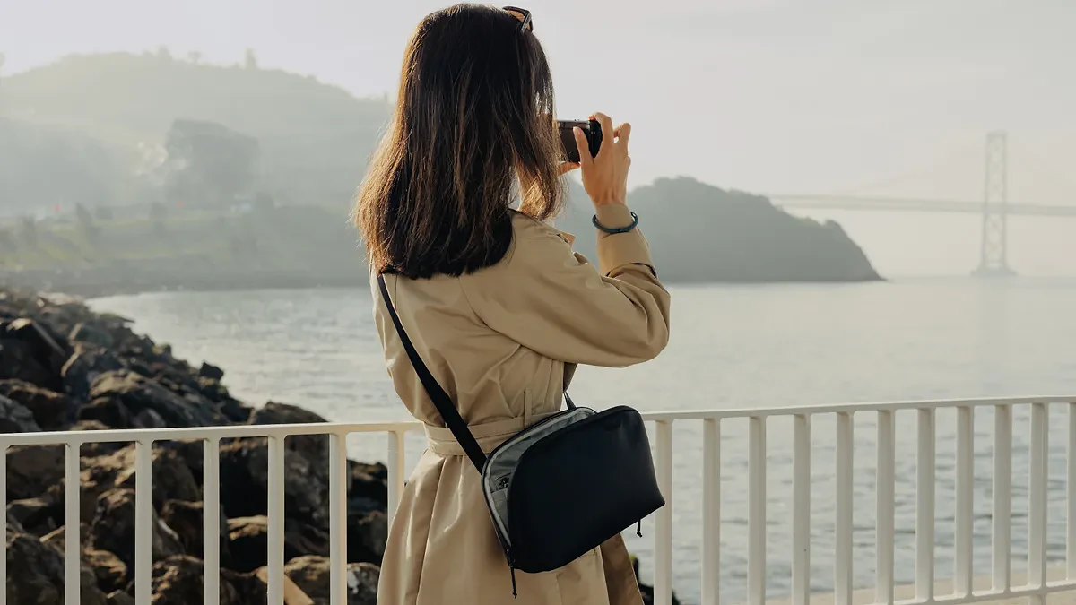 Woman wearing the Peak Design Travel Crossbody 3L bag while taking a photograph of a river and a bridge with a small compact camera