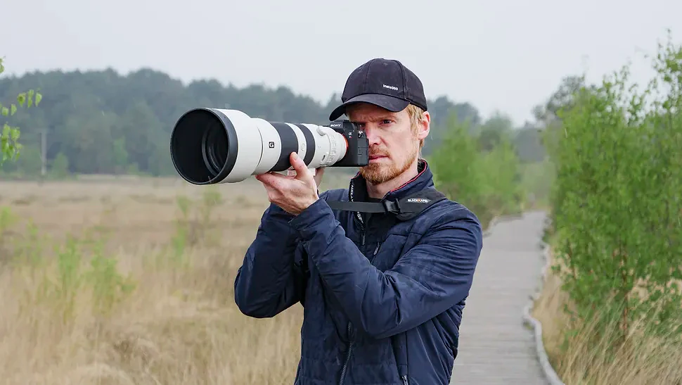 Man holding the Sony A7R VI up to his eye with the FE 400-800mm F6.3-8 G OSS supertelephoto zoom lens attached, he's standing on a boardwalk in a nature reserve