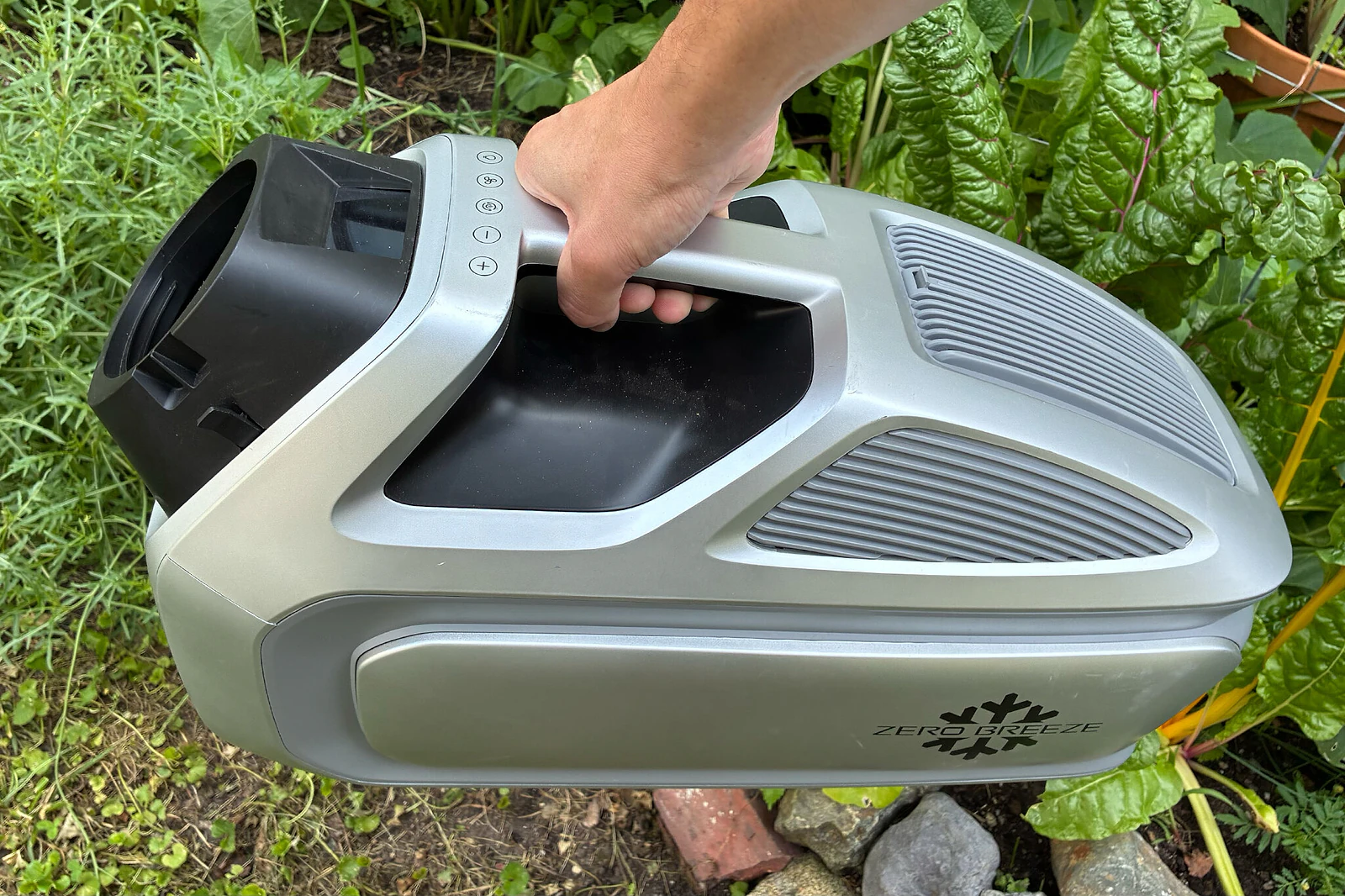 A person's hand holding the Zero Breeze Mark 3 cordless air conditioner in an outdoor environment.
