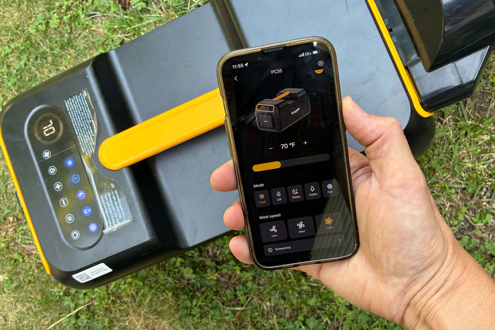 A person's hand holding a smart phone displaying an app for a portable AC, with that same portable AC resting on the grass in the background.