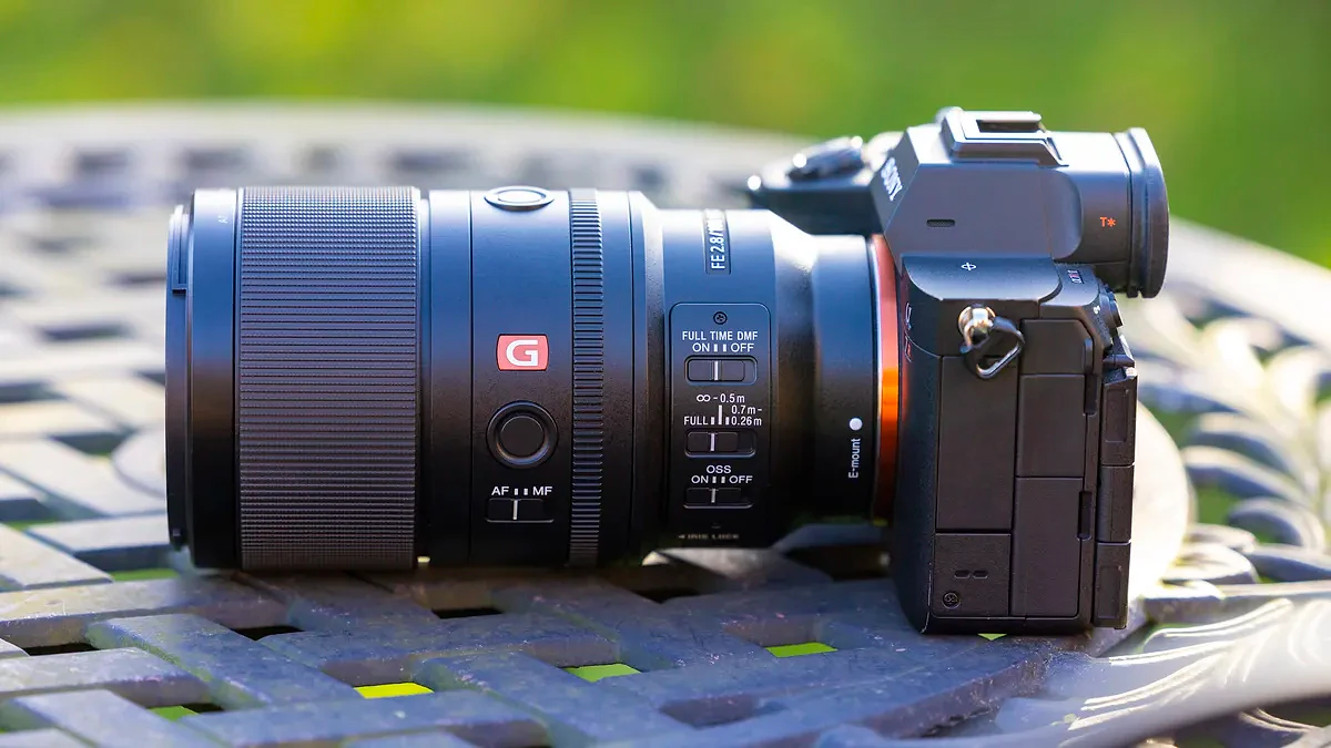 A product shot of the Sony FE 100mm f/2.8 Macro GM OSS attached to a Sony A7R IV outdoors on a metal garden table with a blurry background