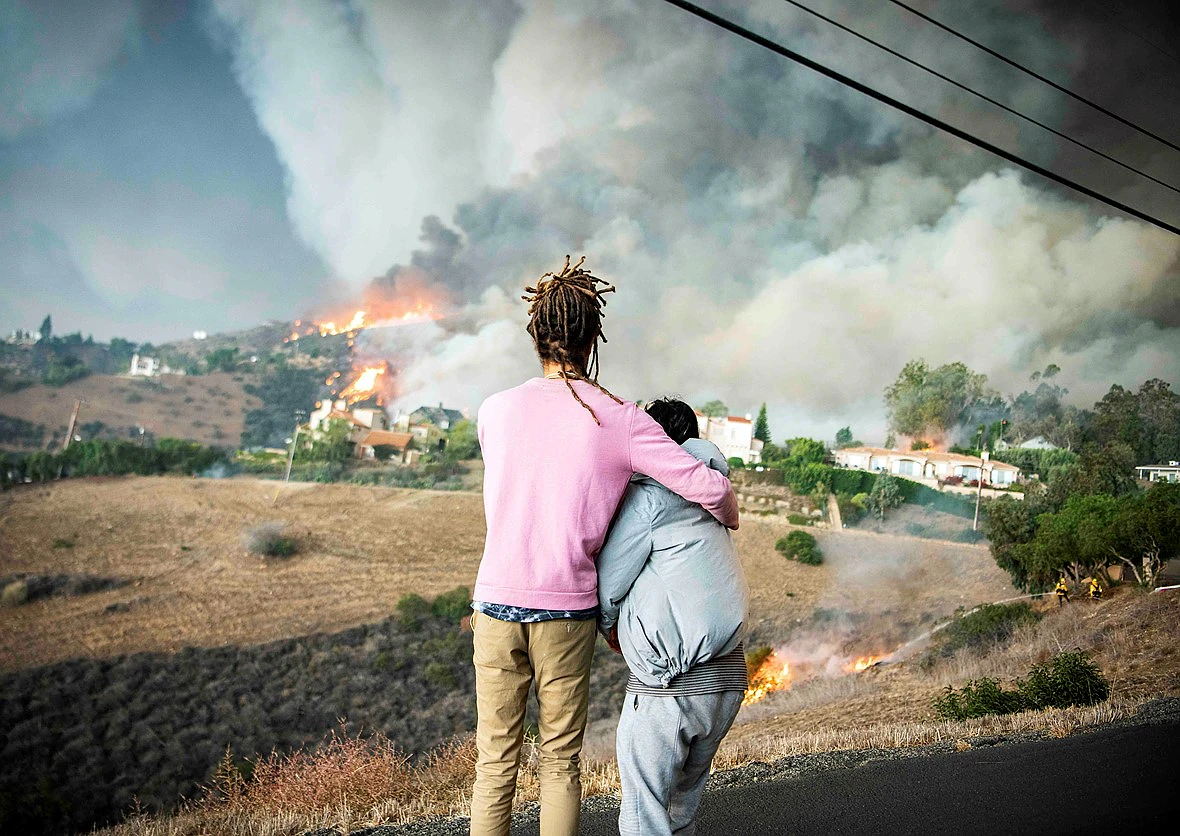 two people stand in an embrace looking out at a large fire engulfing a hillside with houses