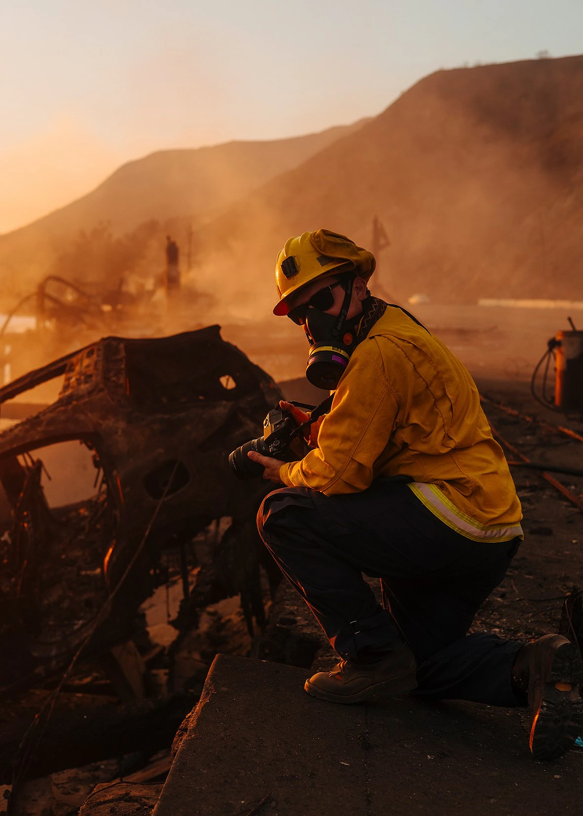 a person wearing a respirator helmet and nomex hacjet kneels down holding a camera amidst charred vehicles and smoke