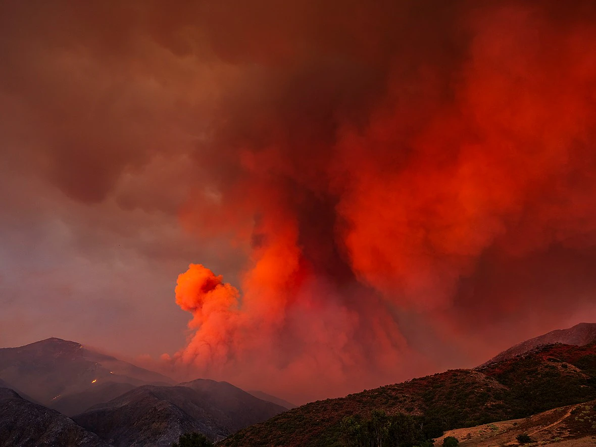 a large plume of red smoke fills the sky above mountains