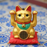A golden Maneki Neko (beckoning cat) figurine with raised paw sits on a red cushion base atop blue a
