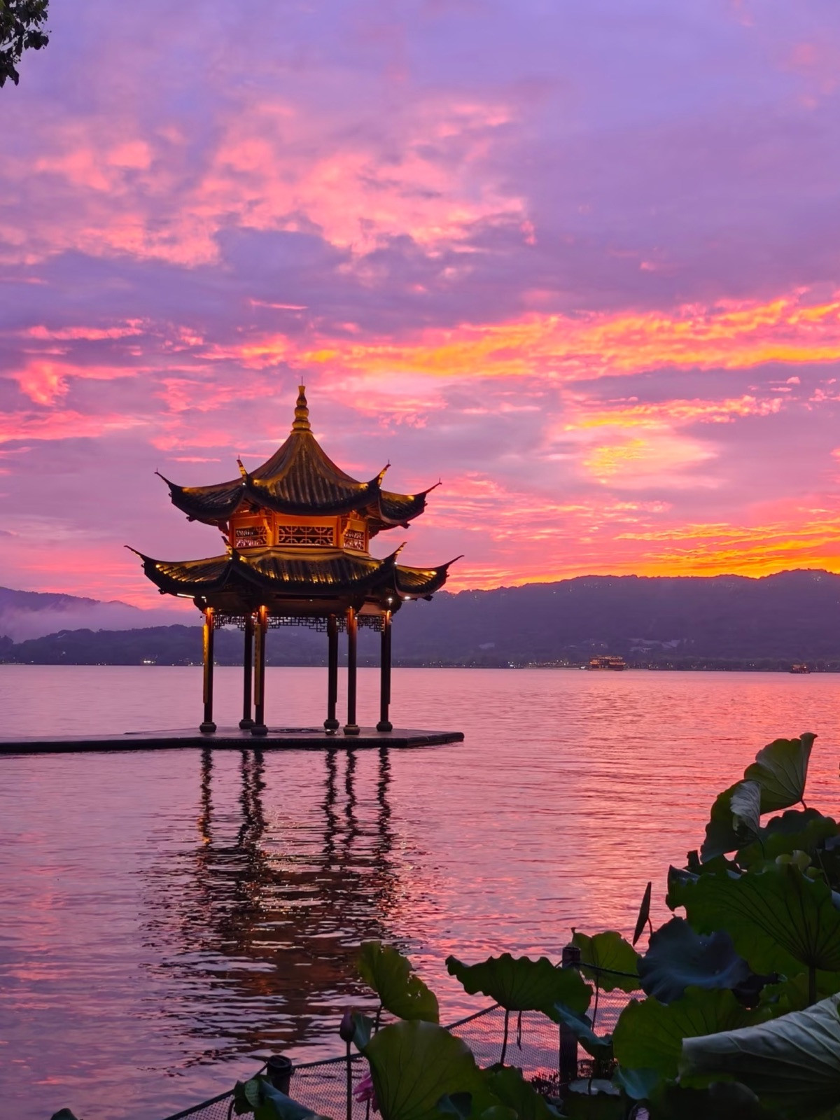 A traditional Chinese pavilion over a lake with lotus leaves in the foreground at sunset