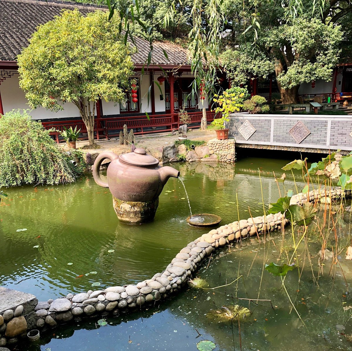 A traditional Chinese garden with a large teapot-shaped fountain pouring water into a koi pond surrounded by trees and a covered walkway