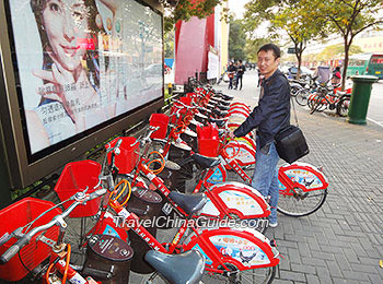 A man standing next to a row of red public bikes at a Hangzhou bike rental kiosk on a city sidewalk