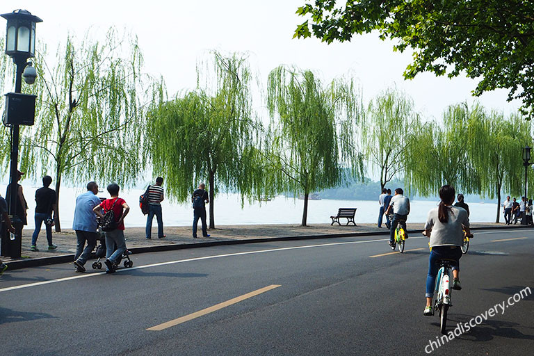 Cyclists and pedestrians along a West Lake causeway with weeping willow trees and a calm lake view