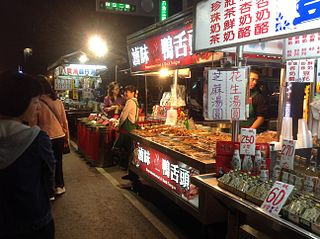 A Hangzhou night market street with food stalls under bright lights, customers browsing skewers and small dishes