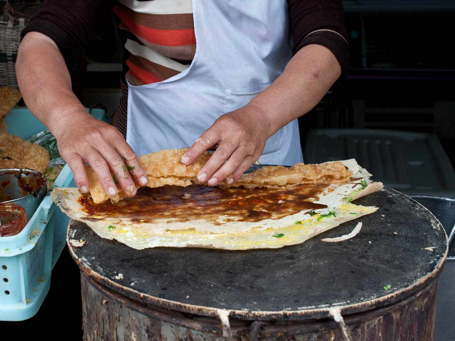 A street vendor making jianbing — hands pressing a crispy youtiao onto a griddled rice crepe smeared with sweet bean sauce