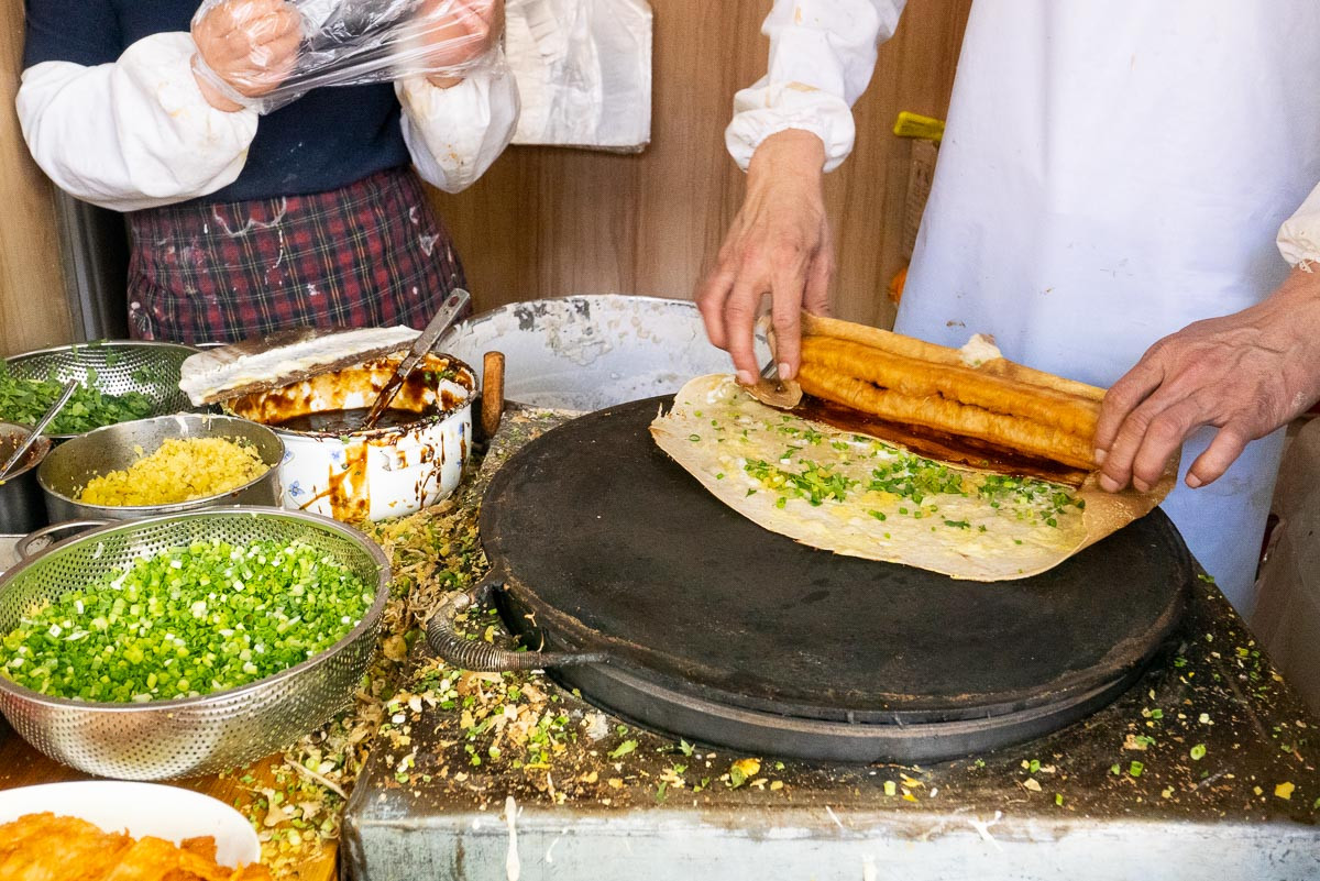 Hands placing a crispy golden youtiao into a freshly griddled jianbing crepe with scallions and cilantro