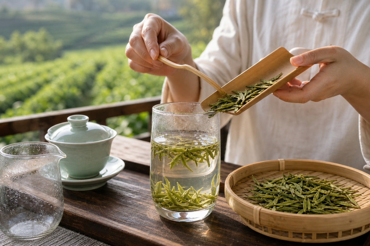 Hands adding fresh Longjing green tea leaves from a bamboo scoop into a glass pitcher with hot water and tea field in background