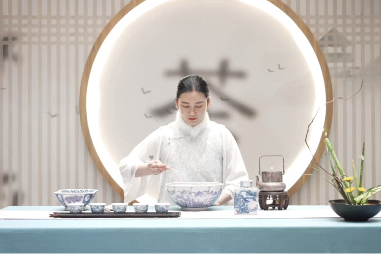 A woman in white traditional dress performing a Chinese tea ceremony with blue-and-white porcelain teaware, framed by a circular wooden opening