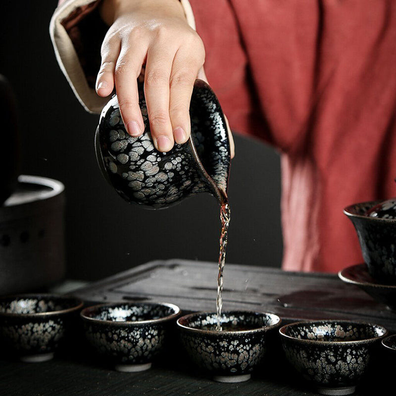 Hands in red traditional clothing pouring tea from a black Jian ware teapot into small black tea cups