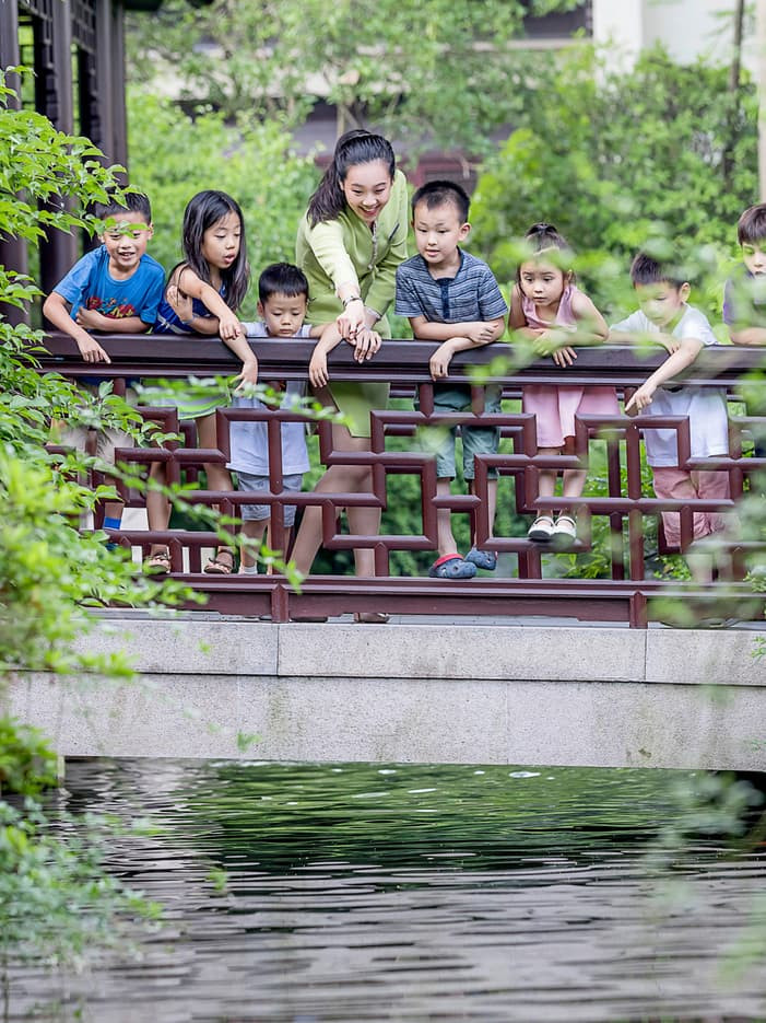 A group of children and a teacher leaning on a wooden bridge railing over water in a classical Chinese garden in Hangzhou