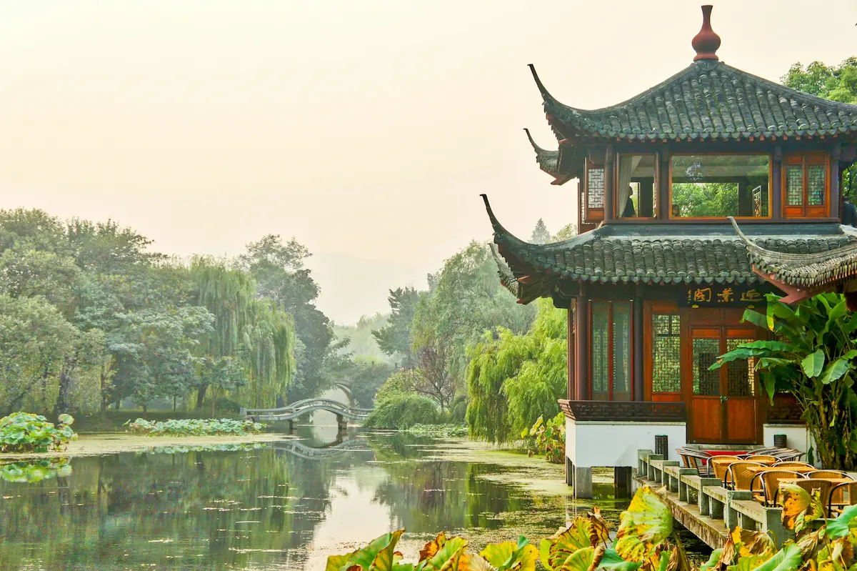 A traditional Chinese pavilion with curved roof beside a lotus pond, misty willows and a stone bridge in the background