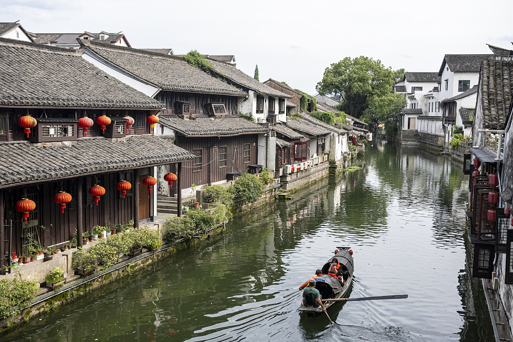 A water town canal in Shaoxing with traditional dark-roofed buildings on both sides, red lanterns, and a small wooden boat with rowers