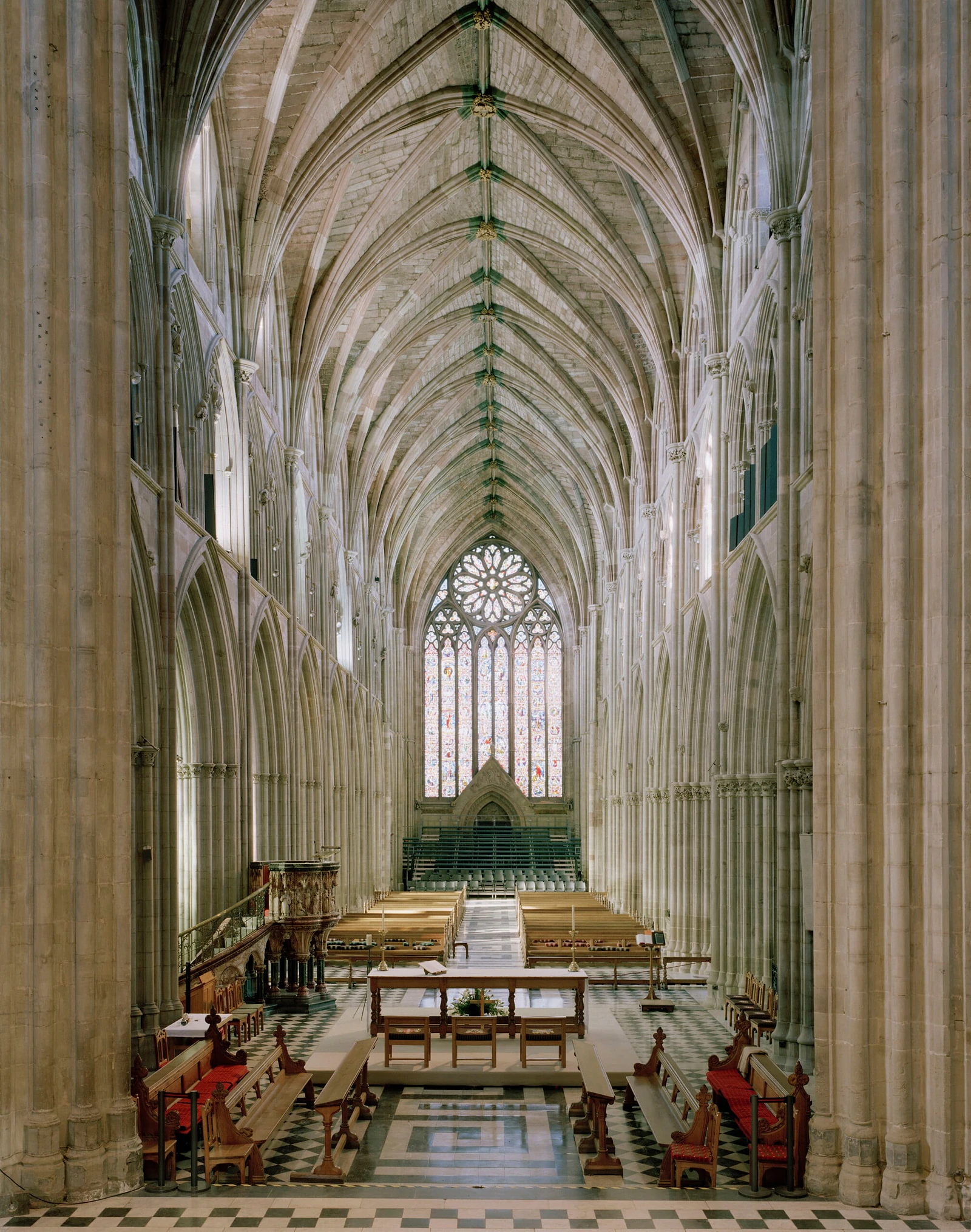 The interior of a Gothic cathedral features a long nave with vaulted ceilings, stone columns, wooden pews, and a stained-glass window at the far end.