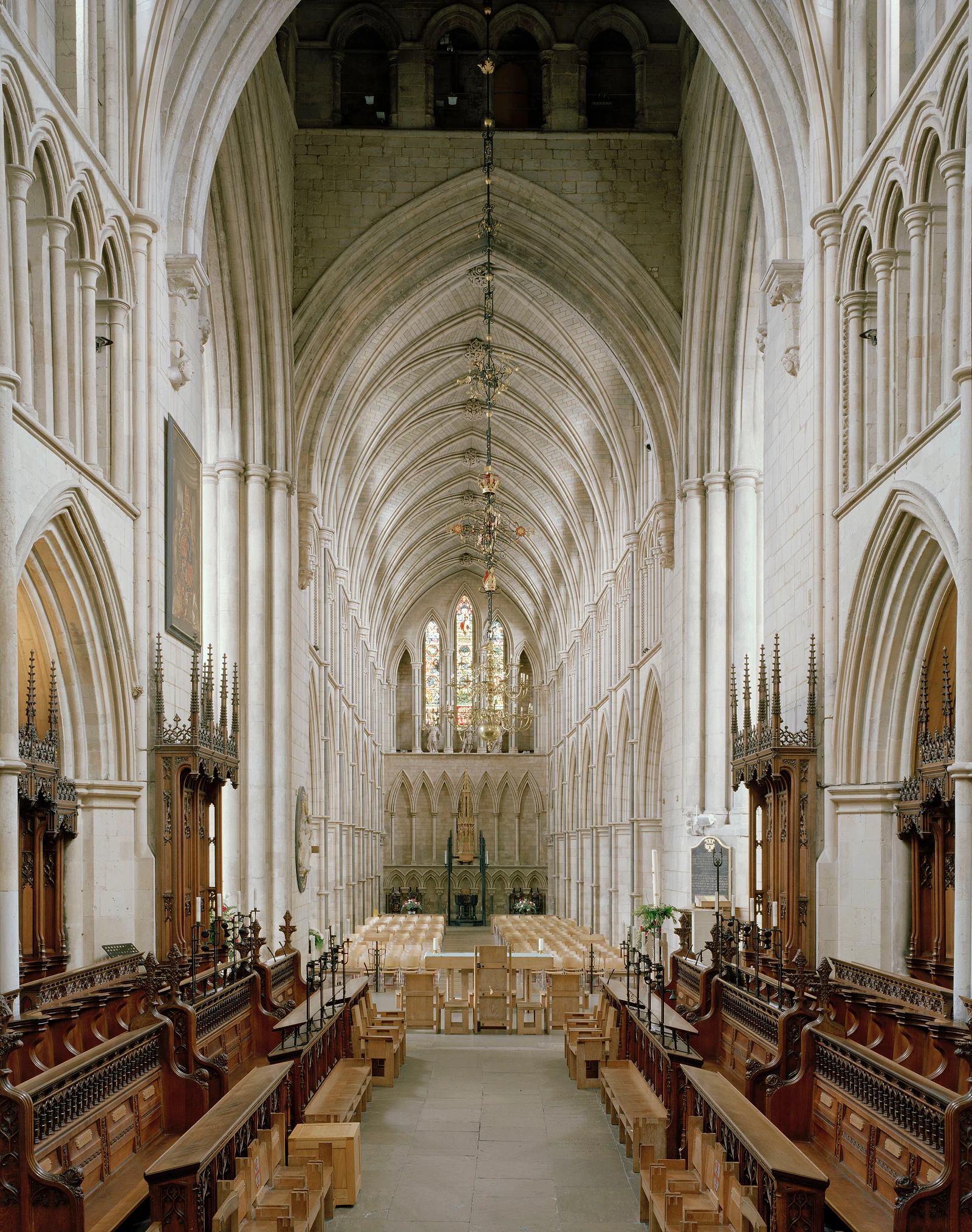 A symmetrical view down the center aisle of a stone cathedral, featuring rows of wooden choir stalls, a high vaulted ceiling, and stained glass windows at the far end.