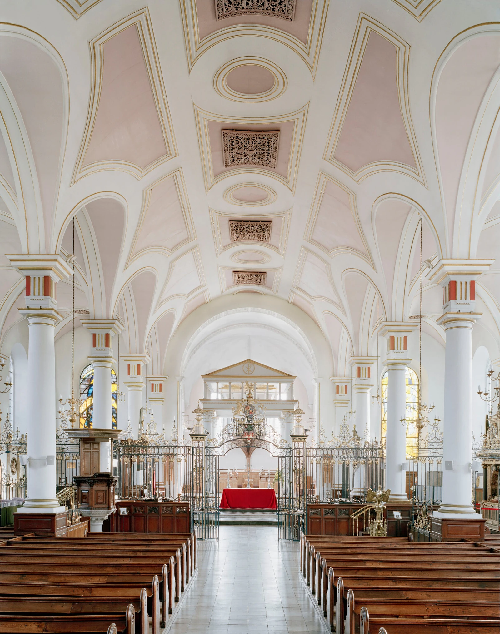The interior of a church features rows of wooden pews leading toward a central altar covered with a red cloth, framed by ornate metal gates and white columns supporting a vaulted ceiling.