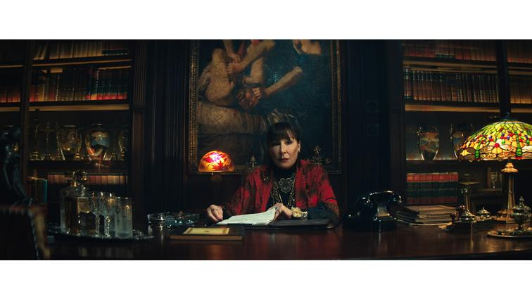 None A woman wearing a black shirt and a red sweater sitting at a massive desk in an office lined with leather-bound books.
