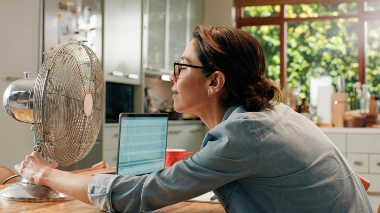 None A woman holds a desk fan in front of her face while sitting at a home desk with a laptop.