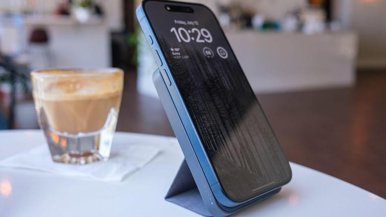 None iPhone connected to a slim portable charger on a stand. They're both on a table in a brightly lit cafe along with a cortado in a glass.