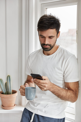 Vertical shot of serious attractive man dressed in casual clothes, spends weekend at home, chats onl.jpg