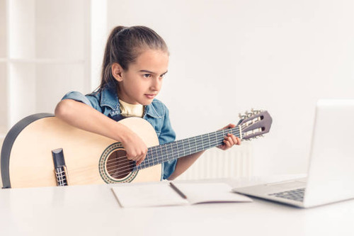 Focused little kid playing acoustic guitar and watching online course on laptop while practicing at .jpg