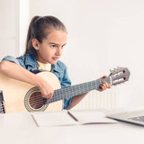 Focused little kid playing acoustic guitar and watching online course on laptop while practicing at 