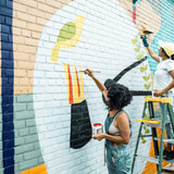 Two Female artists painting large wall mural. They are dressed in casual work clothes. Exterior of p