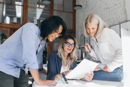 Beautiful blonde girl in glasses showing report to asian colleague which standing beside her table. .jpg
