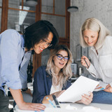Beautiful blonde girl in glasses showing report to asian colleague which standing beside her table. 