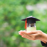 Woman hand holding coins money in glass bottle with graduates hat on natural green background, Savin
