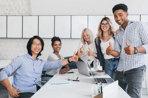 Blonde secretary sitting on table while office workers posing with thumbs up. Indoor portrait of hap.jpg