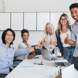 Blonde secretary sitting on table while office workers posing with thumbs up. Indoor portrait of hap