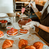 Woman holds spoon with pouring down frosting above bowl at metal table with tasty croissants in craf