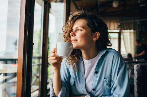 Smiling calm young woman drinking coffee looking out the window sitting at table in restaurant..jpg