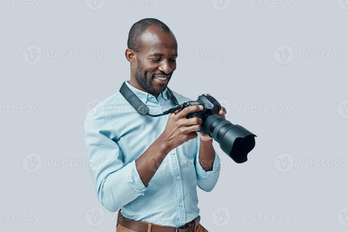 happy young african man using digital camera and smiling while standing against grey background phot.jpg